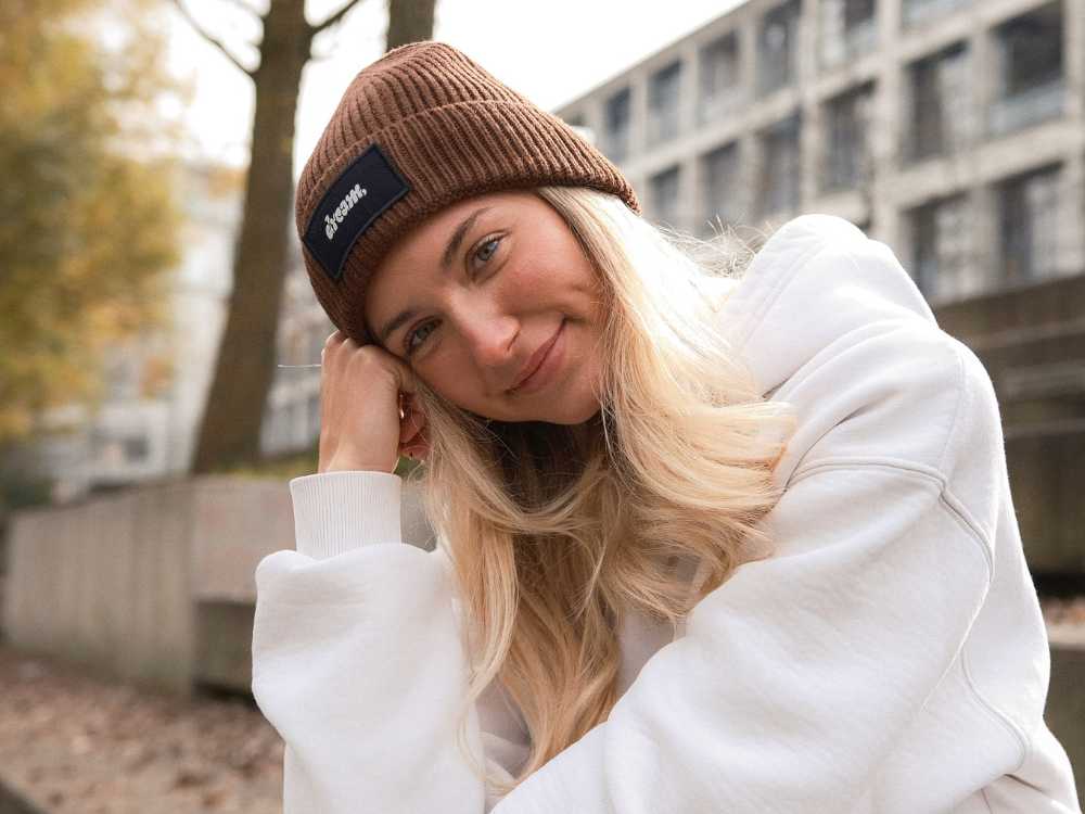 Lächelnde Kundin trägt eine braune, personalisierte Mister Merch Beanie Mütze mit Patch und weißem Logo im urbanen Außenbereich.