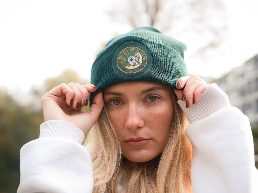 Porträt einer Kundin, die eine dunkelgrüne, personalisierte Mister Merch Beanie mit dem Logo des Schützenvereins "Zum Goldenen Ziel" trägt.