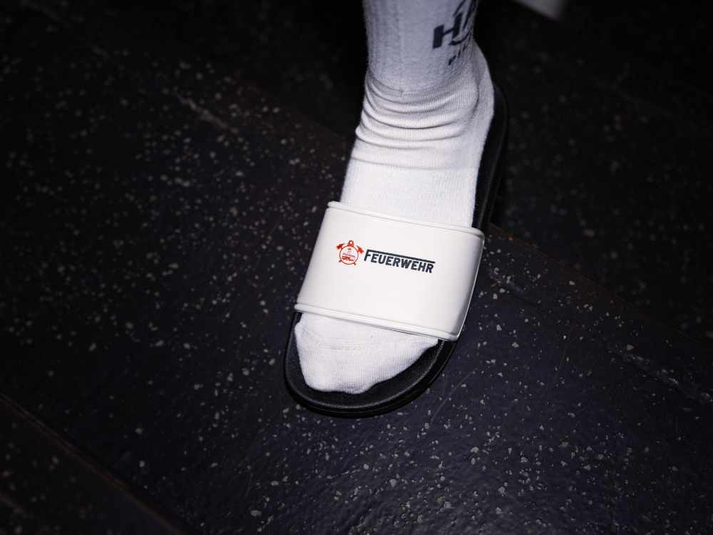 Personalisierte Badeschlappe von Mister Merch in Schwarz-Weiß mit rotem Feuerwehr-Logo, getragen über einer weißen Socke auf dunklem Untergrund.