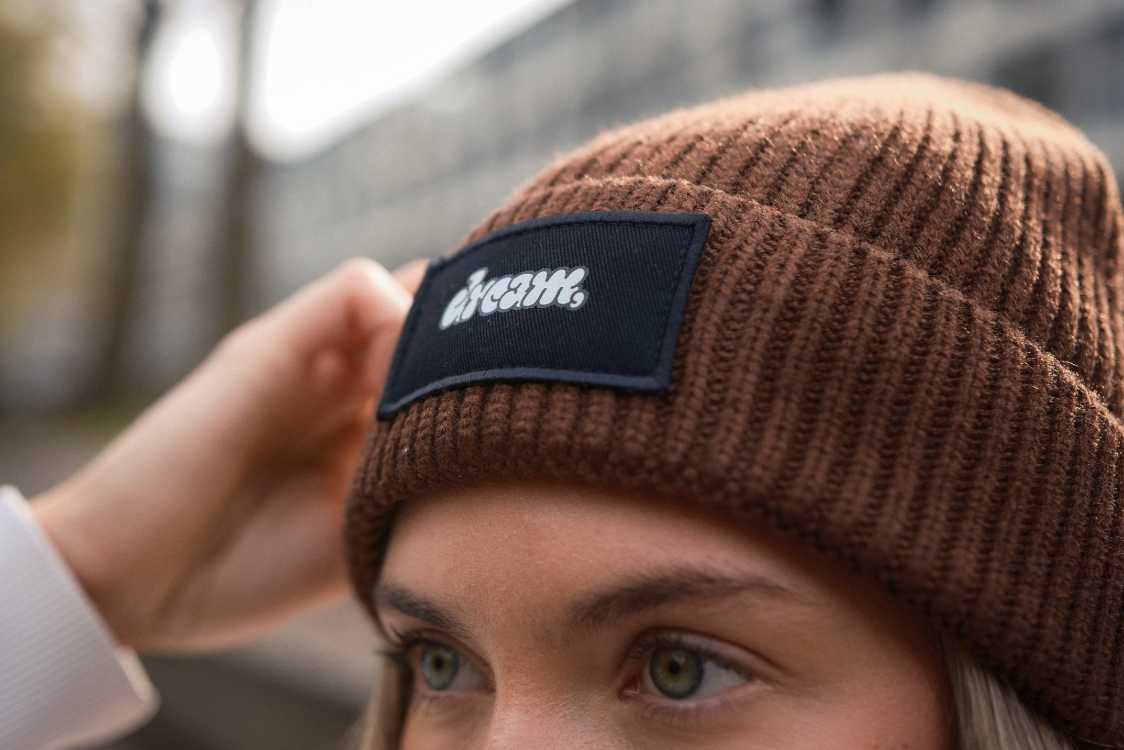 Nahaufnahme einer braunen, personalisierten Strickmütze (Beanie) von Mister Merch mit einem aufgenähten, dunkelblauen Patch, der einen weißen, individuellen Schriftzug zeigt.