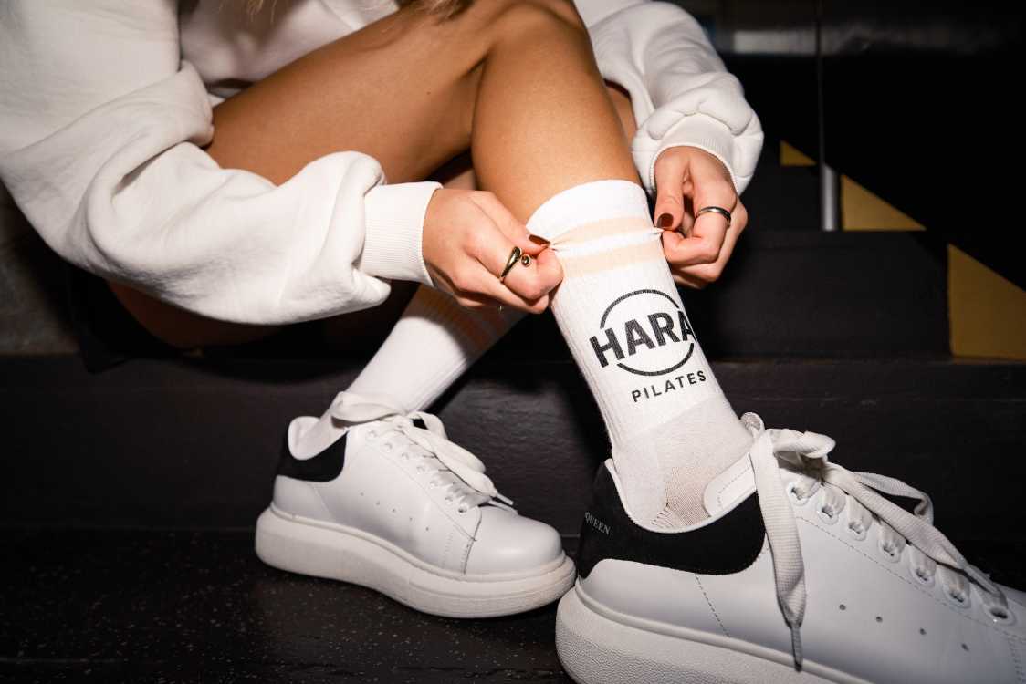 Weiße, personalisierte Socke von Mister Merch mit schwarzem "HARA PILATES" Logo und beigefarbenen Streifen, getragen zu weißen Sneakern.
